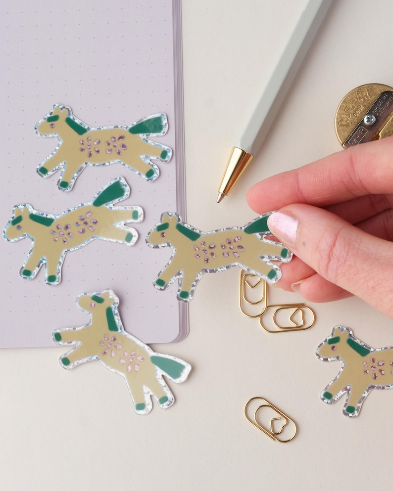 May include: Five gold glitter horse stickers with green and pink details. The stickers are on a white surface with a notebook and a pen in the background.