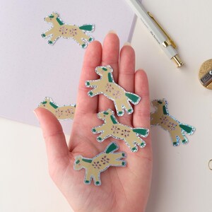 May include: A hand holding six small, glittery horse stickers. The stickers are yellow with green outlines and have a glittery finish. The stickers are on a white background.