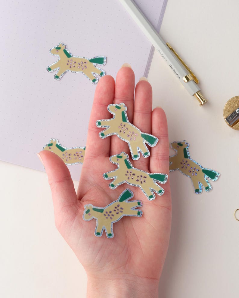 May include: A hand holding six small, glittery horse stickers. The stickers are yellow with green outlines and have a glittery finish. The stickers are on a white background.
