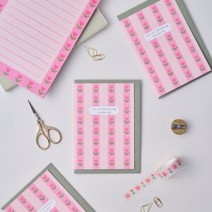 May include: Four pink and green greeting cards with the text "you are blooming wonderful" and a pink floral pattern. The cards are on a white surface with a pencil sharpener, scissors, and washi tape.