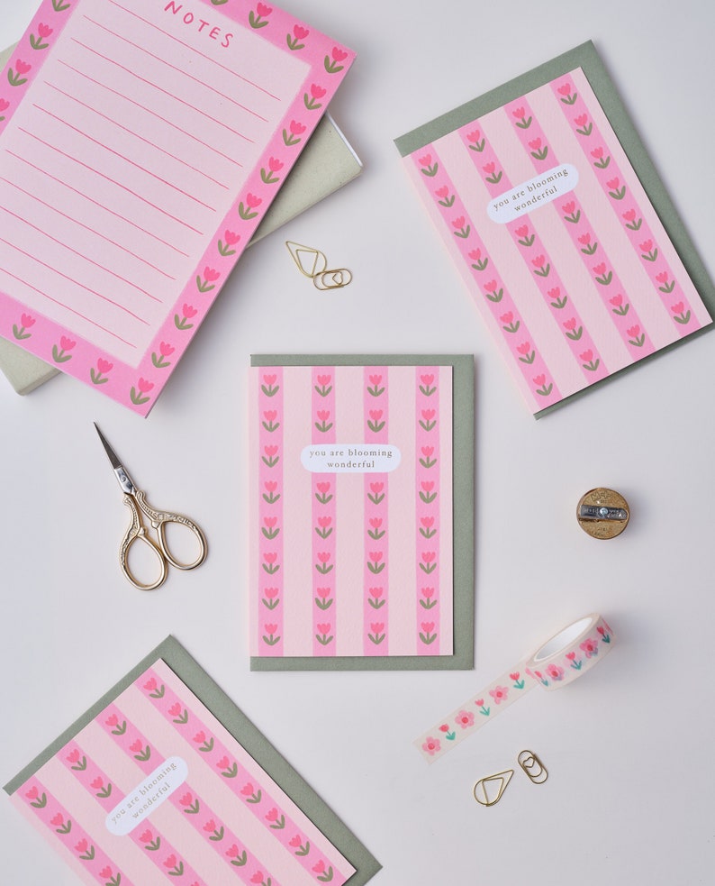 May include: Four pink and green greeting cards with the text "you are blooming wonderful" and a pink floral pattern. The cards are on a white surface with a pencil sharpener, scissors, and washi tape.