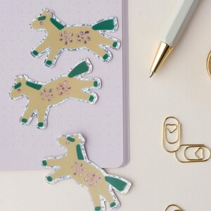 May include: Three yellow horse stickers with green and pink details on a purple notebook. The stickers have a glittery finish.