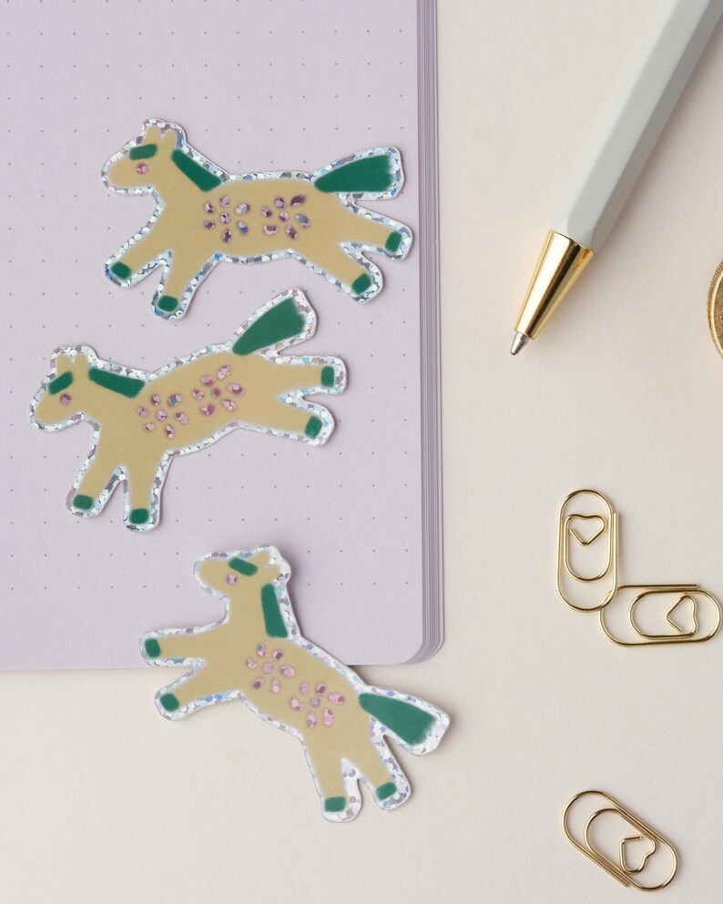 May include: Three yellow horse stickers with green and pink details on a purple notebook. The stickers have a glittery finish.
