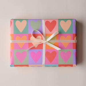 May include: A rectangular gift wrapped in colourful paper with a repeating heart pattern in pink, orange, red, green, and purple. A white ribbon is tied around the gift in a bow.