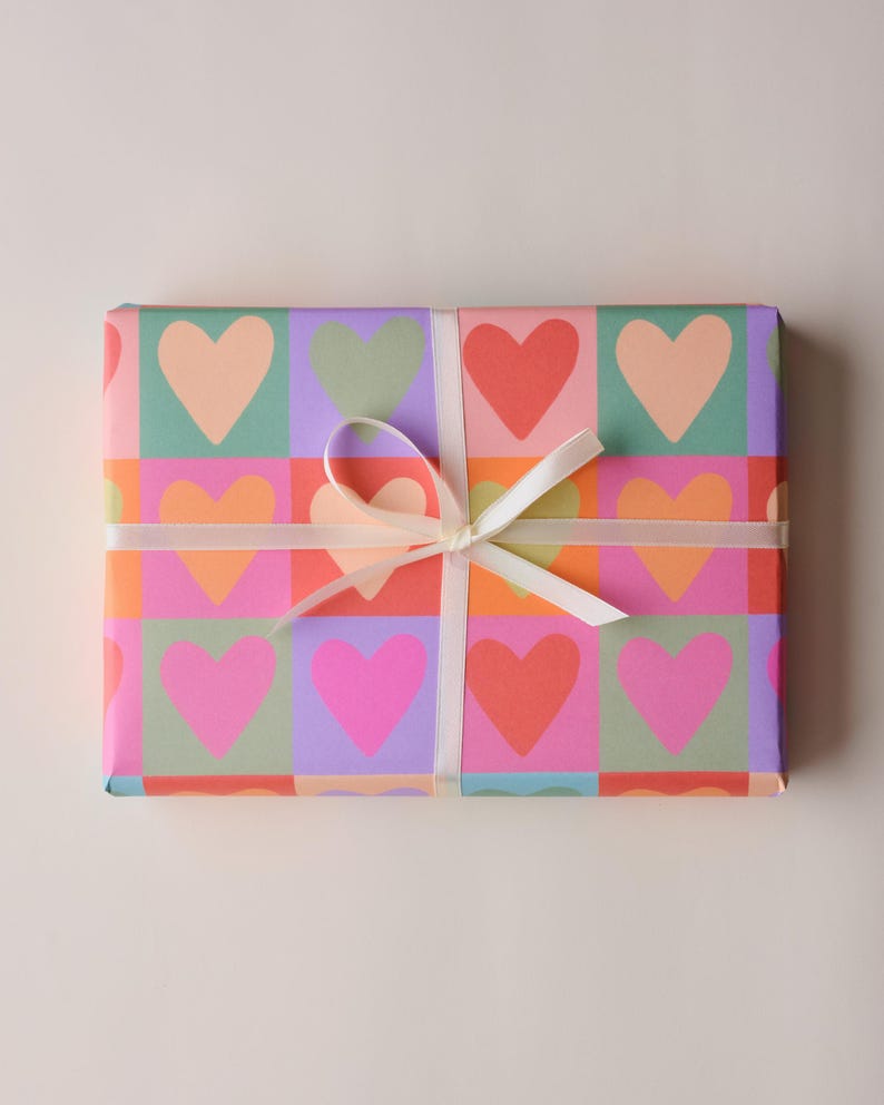 May include: A rectangular gift wrapped in colourful paper with a repeating heart pattern in pink, orange, red, green, and purple. A white ribbon is tied around the gift in a bow.