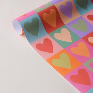 May include: Rolled sheet of wrapping paper featuring a repeating pattern of hearts in squares. The hearts are in shades of green, orange, red, pink, and purple. The background squares are in pink, blue, and orange.