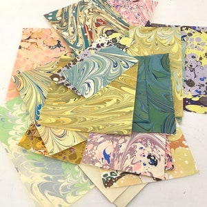 May include: A selection of marbled paper sheets in diverse colours and patterns. The sheets feature swirls of blue, green, yellow, pink, and purple, creating a unique artistic look. The papers are arranged in a layered, overlapping composition.