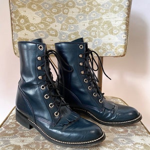 May include: A pair of dark blue leather lace-up boots with fringe detailing on the toe. The boots have a stacked heel and a rubber sole.