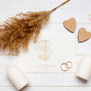 May include: A white card with a gold monogram of the letters "B" and "J" intertwined. The card is on a white wooden surface with a dried grass bouquet, two wooden hearts, and two white candles.