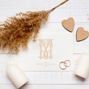 May include: A white card with a gold monogram design of the letters "M" and "V" intertwined. The card is on a white wooden surface with dried pampas grass, two wooden hearts, and two white candles.