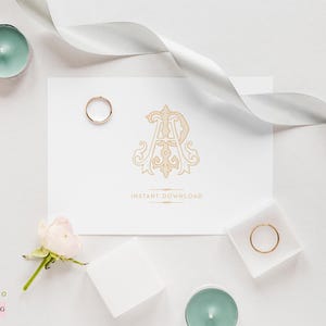 May include: A white card with a gold monogram design featuring the letters "A" and "R" in a decorative font. The card is surrounded by white ribbon, a white rose, and two gold rings.