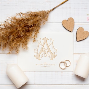 May include: A white card with a gold monogram of the letters "A" and "M" surrounded by decorative flourishes. The card is on a white wooden surface with two wooden hearts, a bundle of dried grass, and two white candles.