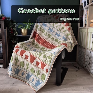 Op de afbeelding: Een gehaakte deken met een herhalend patroon van rode, groene en blauwe motieven, waaronder bomen, cactussen en theepotten. De deken is over een stoel gedrapeerd. De tekst "Crochet pattern" en "English PDF" zijn zichtbaar.