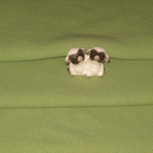 May include: Two small white ceramic sheep figurines with black eyes and noses, sitting on a green fabric surface.
