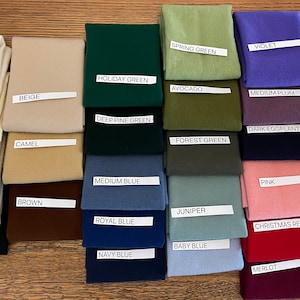 May include: A collection of fabric swatches in various colors, including white, beige, green, blue, pink, and brown. Each swatch has a label with the color name, such as "White", "Holiday Green", and "Pink". The swatches are neatly arranged on a wooden surface.