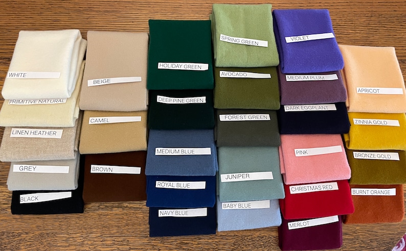 May include: A collection of fabric swatches in various colors, including white, beige, green, blue, pink, and brown. Each swatch has a label with the color name, such as "White", "Holiday Green", and "Pink". The swatches are neatly arranged on a wooden surface.