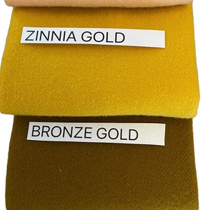 May include: A stack of four fabric swatches in shades of apricot, zinnia gold, bronze gold, and burnt orange. Each swatch has a label with its color name. The fabrics appear to be a soft, textured material.