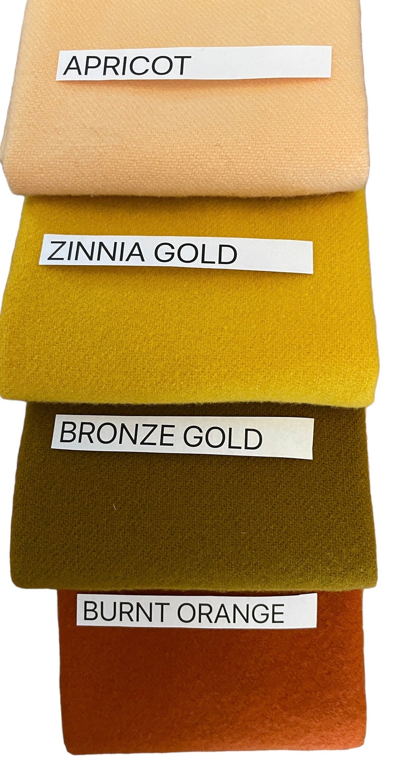 May include: A stack of four fabric swatches in shades of apricot, zinnia gold, bronze gold, and burnt orange. Each swatch has a label with its color name. The fabrics appear to be a soft, textured material.