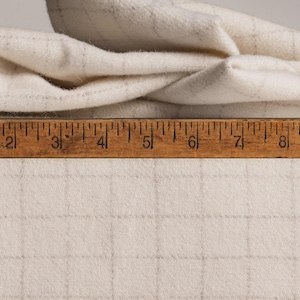 May include: Cream-colored fabric with a grid pattern and a wooden ruler. The fabric is folded over, showing its texture. The ruler is marked in inches, providing a size reference for the material.