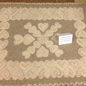 May include: A square rug featuring a beige background with a cream-colored heart pattern. The border consists of a repeating heart design, while the center displays a cross-shaped arrangement of hearts. A small white card reads Have A Heart, Core Wool, Dark Gold.