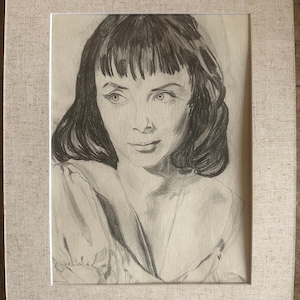 May include: A graphite pencil portrait drawing of a person with shoulder-length hair and bangs, framed in a light beige textured mount. The artwork captures the facial features, including defined eyes and a soft expression, rendered in shades of grey.