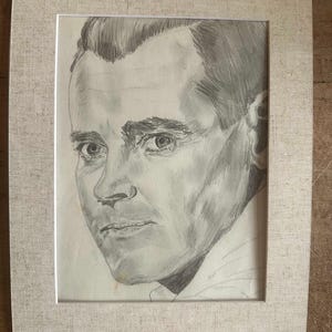 Op de afbeelding: Een grafietpotloodtekening van een portret van een persoon met kort haar, weergegeven in grijstinten op wit papier. Het kunstwerk is ingelijst in een lichtbeige, getextureerde passe-partout, wat de gedetailleerde schaduwwerking van het gezicht benadrukt.
