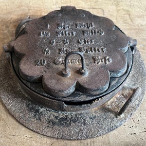 May include: Antique cast iron waffle iron with a floral design. The iron is dark brown with a handle and a hinged lid. The lid has embossed text. The waffle iron is sitting on a wooden surface.