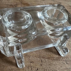 Könnte beinhalten: Ein doppelter Salzstreuer aus klarem Glas mit zwei runden Vertiefungen und einem rechteckigen Sockel. Der Salzstreuer hat zwei kleine Füße und besteht aus dickem, klarem Glas.