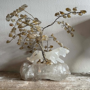 Pode incluir: Uma escultura decorativa de árvore de cristal com uma figura de tigre branco. A árvore tem uma estrutura de arame com folhas de cristal âmbar. O tigre é esculpido em uma pedra branca translúcida e assenta sobre uma grande base de cristal transparente.