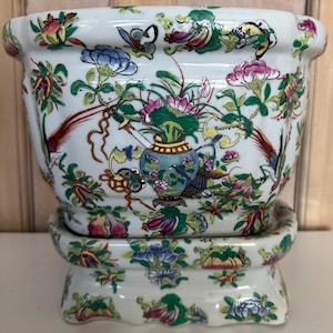 Könnte beinhalten: Ein weißer Keramik-Blumentopf mit passendem Sockel, verziert mit farbenfrohen Blumen- und Vogelmotiven. Der Topf hat eine breite, abgerundete Form und ist mit rosa, blauen und grünen Blumen und einer blauen Vase verziert. Der Sockel ist ebenfalls mit Blumen verziert.