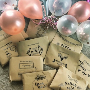 May include: A collection of brown paper envelopes with different messages written on them. The envelopes are decorated with black ink drawings and are surrounded by pink and clear balloons.