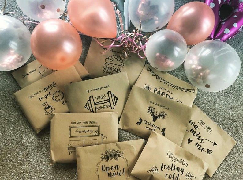 May include: A collection of brown paper envelopes with different messages written on them. The envelopes are decorated with black ink drawings and are surrounded by pink and clear balloons.