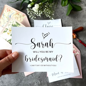 May include: A white card with black text that says "Sarah, will you be my bridesmaid? I can't say I do without you!" with a small heart drawing above the name.