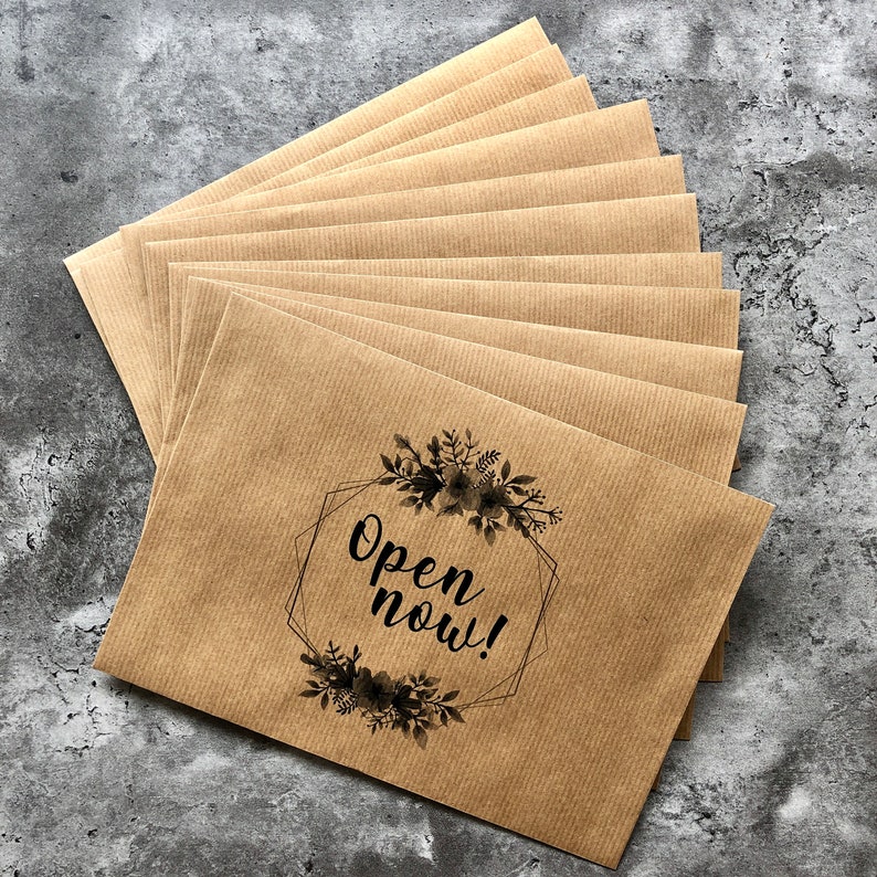 May include: A stack of brown kraft paper envelopes with a black floral design and the text "Open now!" printed on the front.