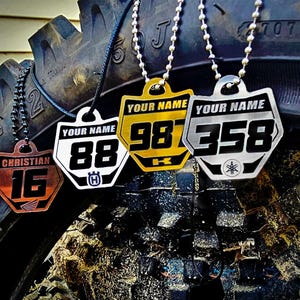 May include: Four different metal necklaces with personalised racing numbers and names. The necklaces are hanging from a tyre. The numbers are 16, 88, 98, and 358. The names are Christian, Your Name, Your Name, and Your Name.