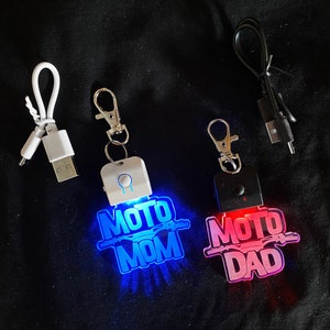May include: Two keychains with light-up designs. The keychain on the left has a blue light and says "Moto Mom". The keychain on the right has a pink light and says "Moto Dad".