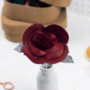 May include: A burgundy leather rose with gray felt leaves in a small white vase. The handmade flower is the focal point, with a craft box and sewing supplies in the background. The rose is a decorative item.