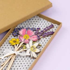 Op de afbeelding: Een set houten bloemensjablonen in een bruine doos. De bloemen zijn geschilderd in tinten roze, geel, wit, paars en groen. De doos is bekleed met wit papier met zwarte stippen.