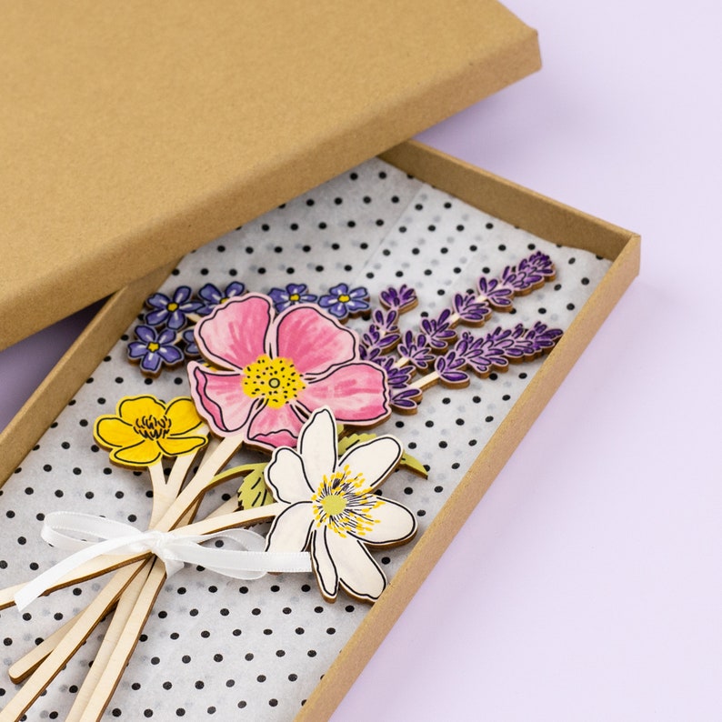 Op de afbeelding: Een set houten bloemensjablonen in een bruine doos. De bloemen zijn geschilderd in tinten roze, geel, wit, paars en groen. De doos is bekleed met wit papier met zwarte stippen.