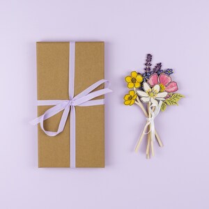 Op de afbeelding: Een bruine cadeaudoos met een lavendelkleurig lint dat in een strik is gebonden. De doos staat naast een boeket papierbloemen in tinten geel, roze, paars en groen.