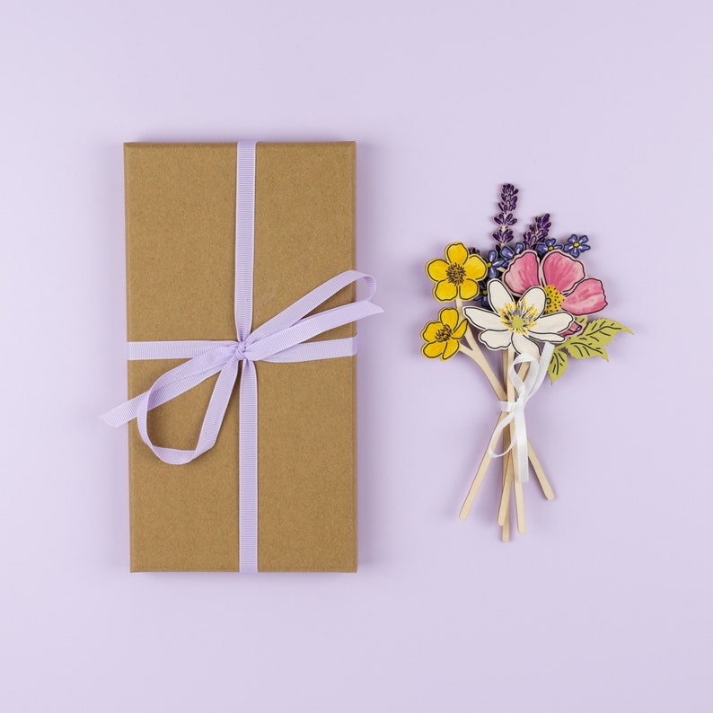 Op de afbeelding: Een bruine cadeaudoos met een lavendelkleurig lint dat in een strik is gebonden. De doos staat naast een boeket papierbloemen in tinten geel, roze, paars en groen.