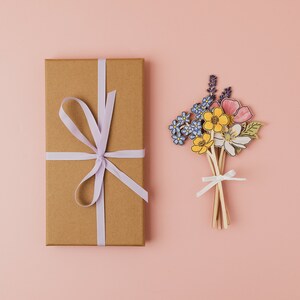 Op de afbeelding: Een bruine cadeaudoos met een lichtpaars lint dat in een strik is gebonden. De doos staat op een roze oppervlak. Naast de doos staat een boeket papierbloemen in tinten blauw, geel en roze.