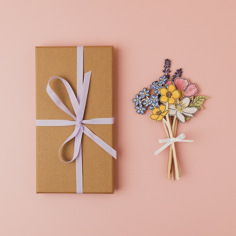 Op de afbeelding: Een bruine cadeaudoos met een lichtpaars lint dat in een strik is gebonden. De doos staat op een roze oppervlak. Naast de doos staat een boeket papierbloemen in tinten blauw, geel en roze.