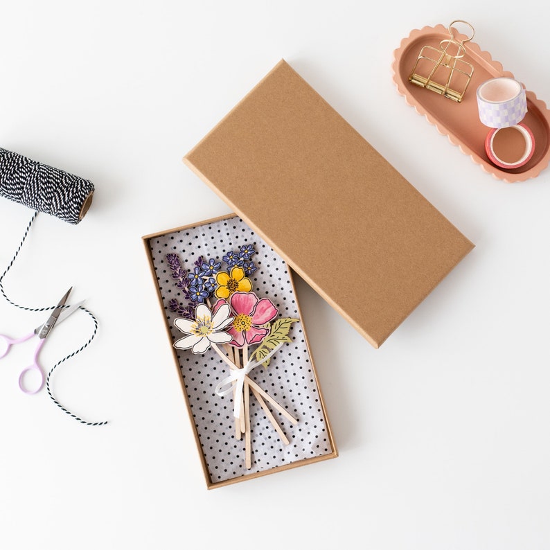 Op de afbeelding: Een bruine kartonnen doos met een witte en zwarte stippen-papierbekleding. De doos bevat een boeket papierbloemen met roze, gele en paarse bloemen. De bloemen zitten op houten stokjes en zijn met een wit lint aan elkaar gebonden.