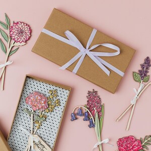 May include: A set of six laser-cut wooden flower cards with a pink background. The cards feature different types of flowers, including lilies, hyacinths, and carnations. The cards are tied with white ribbon and have a "Happy Mother's Day" tag.