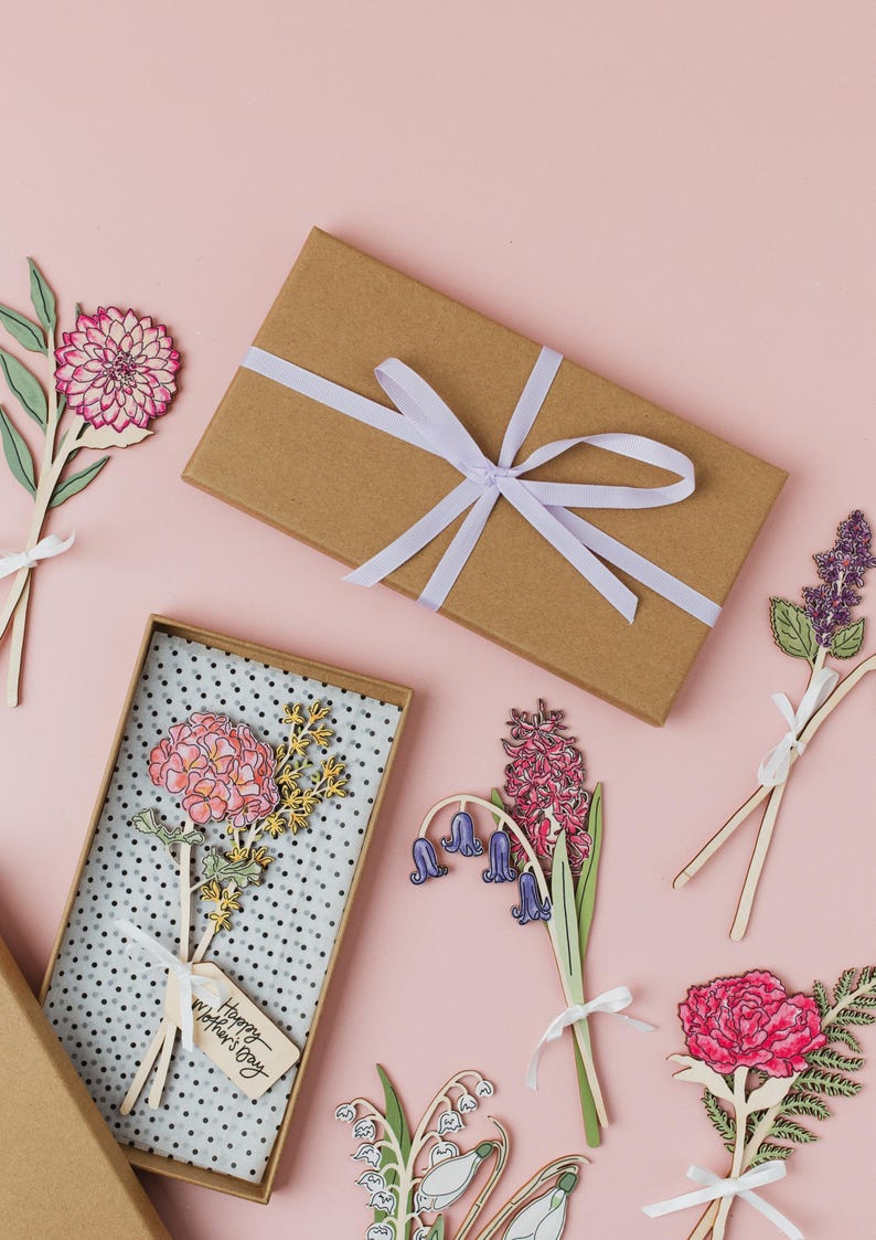 May include: A set of six laser-cut wooden flower cards with a pink background. The cards feature different types of flowers, including lilies, hyacinths, and carnations. The cards are tied with white ribbon and have a "Happy Mother's Day" tag.
