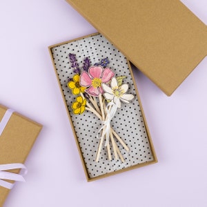 Op de afbeelding: Een bruine doos met een witte stippenpapier voering. In de doos zit een boeket papierbloemen met een wit lint. De bloemen zijn roze, geel, paars en wit.