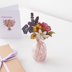Op de afbeelding: Een roze glazen vaas met een boeket papierbloemen. De bloemen zijn geel, roze, paars en blauw. De vaas heeft een getextureerd oppervlak.