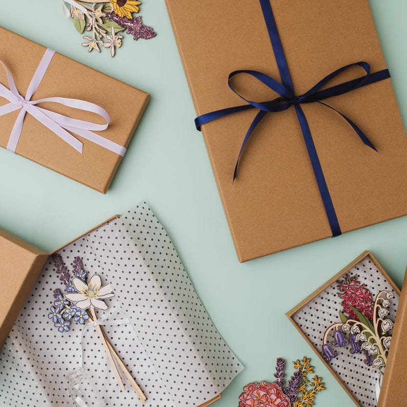 May include: Three brown cardboard gift boxes with blue and purple ribbon. The boxes are on a light blue background. The boxes are decorated with paper flowers and tissue paper.