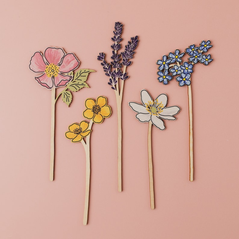 Op de afbeelding: Vijf houten bloemuitsparingen op een roze achtergrond. De bloemen zijn een roze roos, een witte bloem met zes bloemblaadjes, een gele bloem met vier bloemblaadjes, een gele bloem met vijf bloemblaadjes en een tros blauwe vergeet-mij-nietjes.
